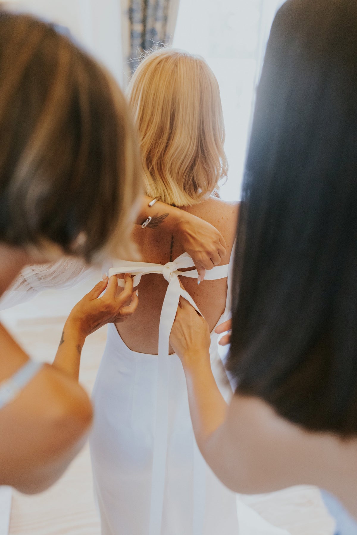 A bride in a minimal wedding dress is being helped by friends to tie the back ribbon, highlighting the elegance of minimal wedding jewelry guide.