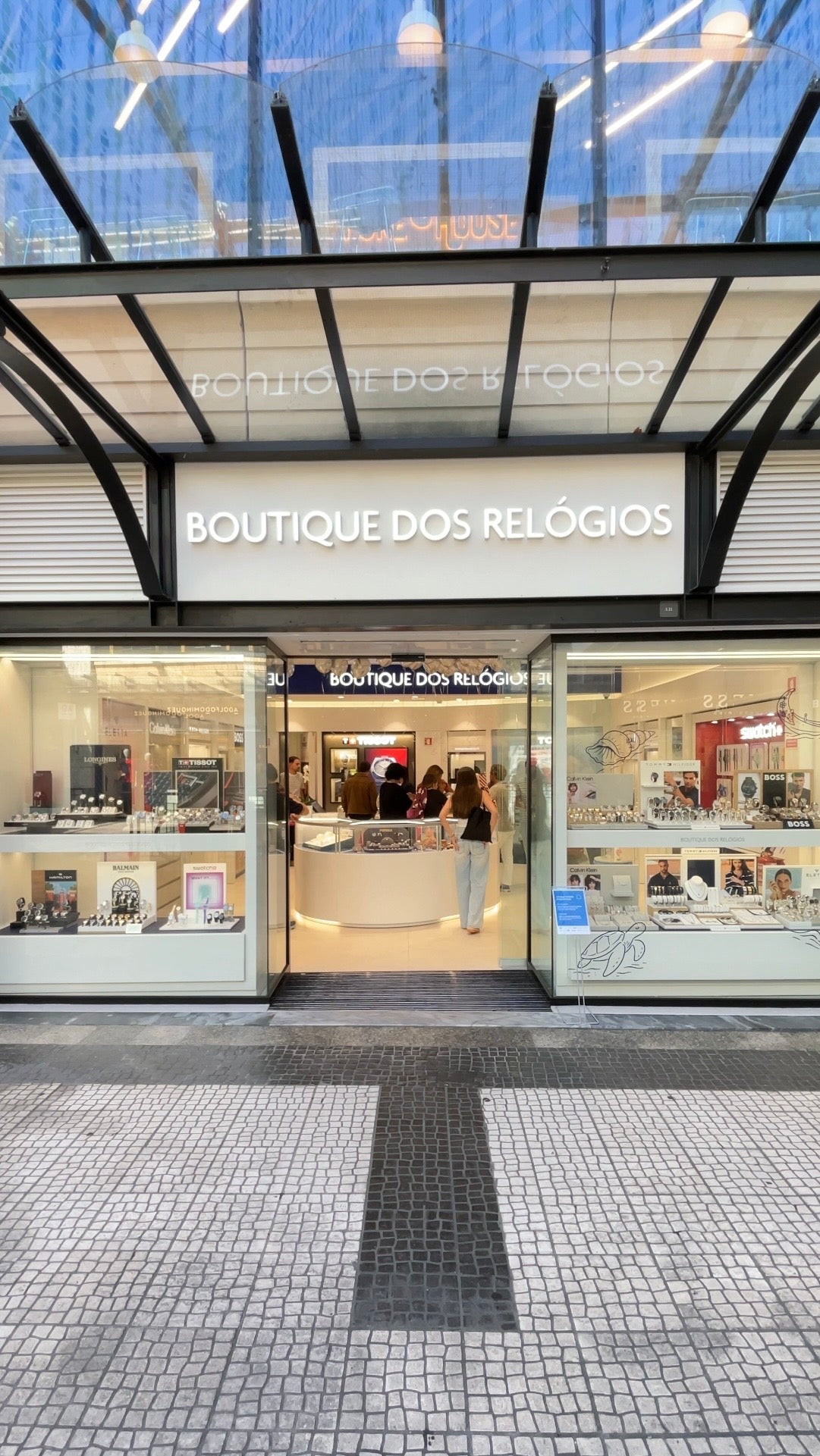 Front exterior of a retail boutique with the sign 'BOUTIQUE DOS RELÓGIOS' above glass display windows and doors. The storefront is lit, with watch displays inside and a tiled walkway in front.