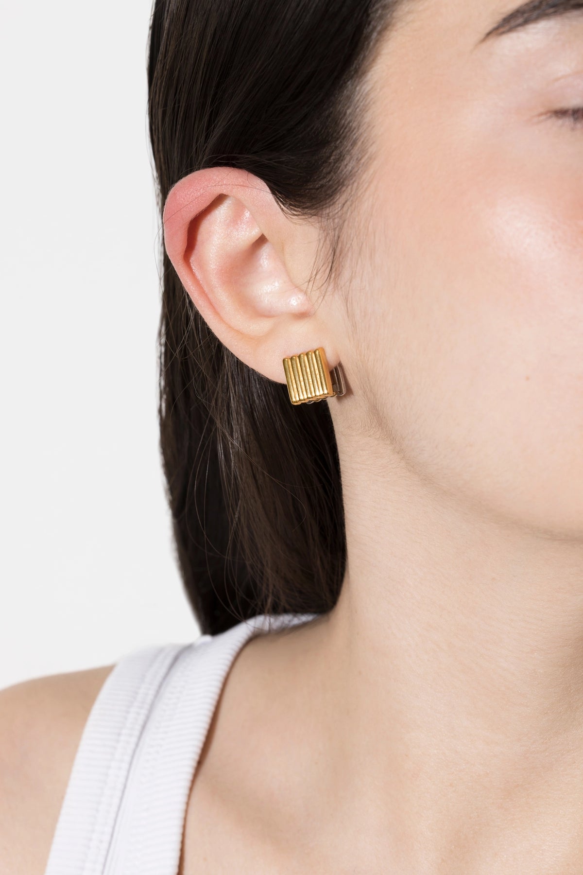 Model's ear wearing a gold rectangular stud earring with vertical ridges, set against dark hair and a light neutral background. CINCO brand jewelry cannelé mini two tones. CINCO brand jewelry cannelé mini two tones