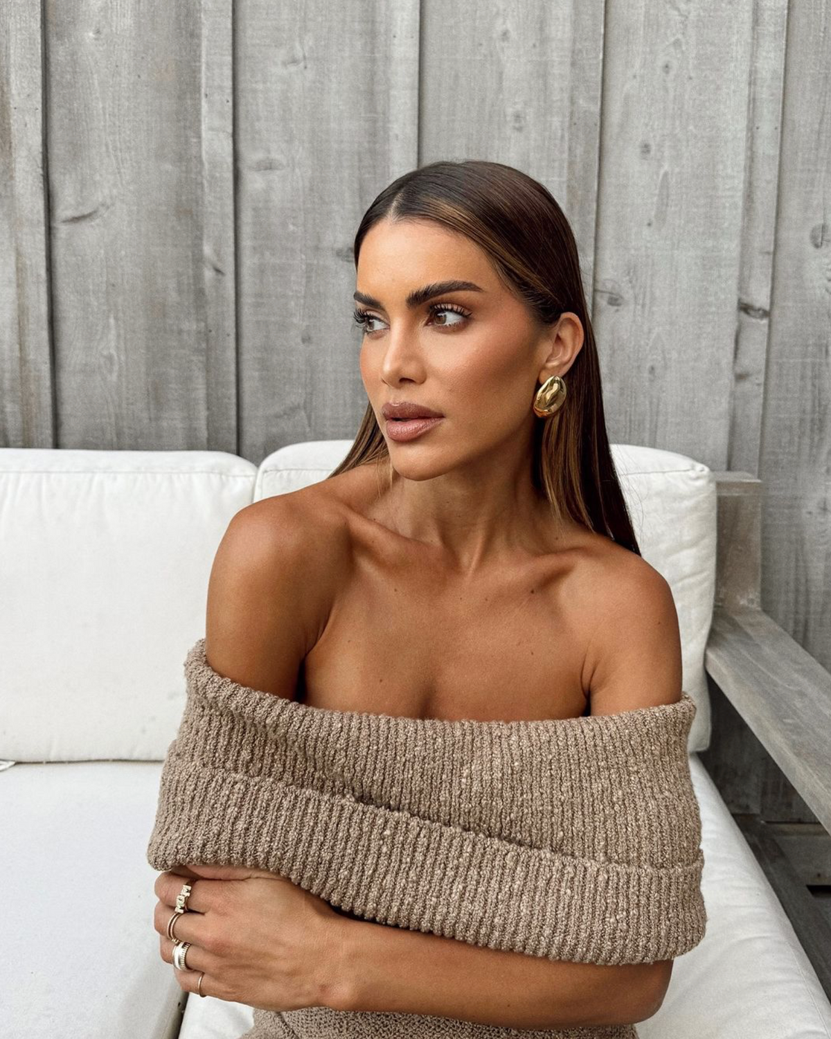 Camila Coelho jewelry collection featuring gold earrings, model wearing a beige off-shoulder sweater against a wooden background.