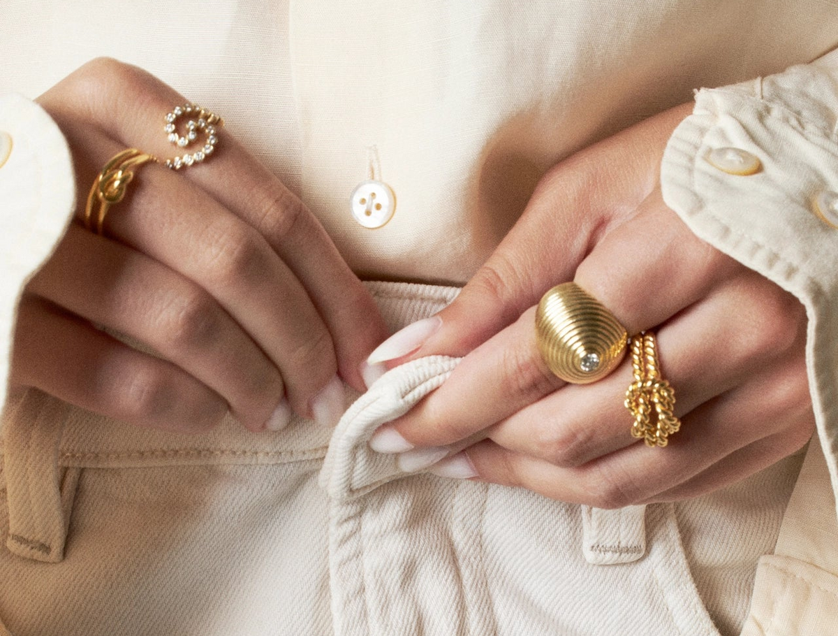 Close-up of a person's hands wearing multiple gold rings, including a spiral ring with clear stones, a textured dome ring, and a rope-knot ring, styled with a cream-colored shirt and trousers.