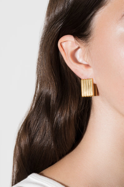 A close-up of a model's ear wearing a gold rectangular stud earring with vertical ridges, set against a clean light gray background.
