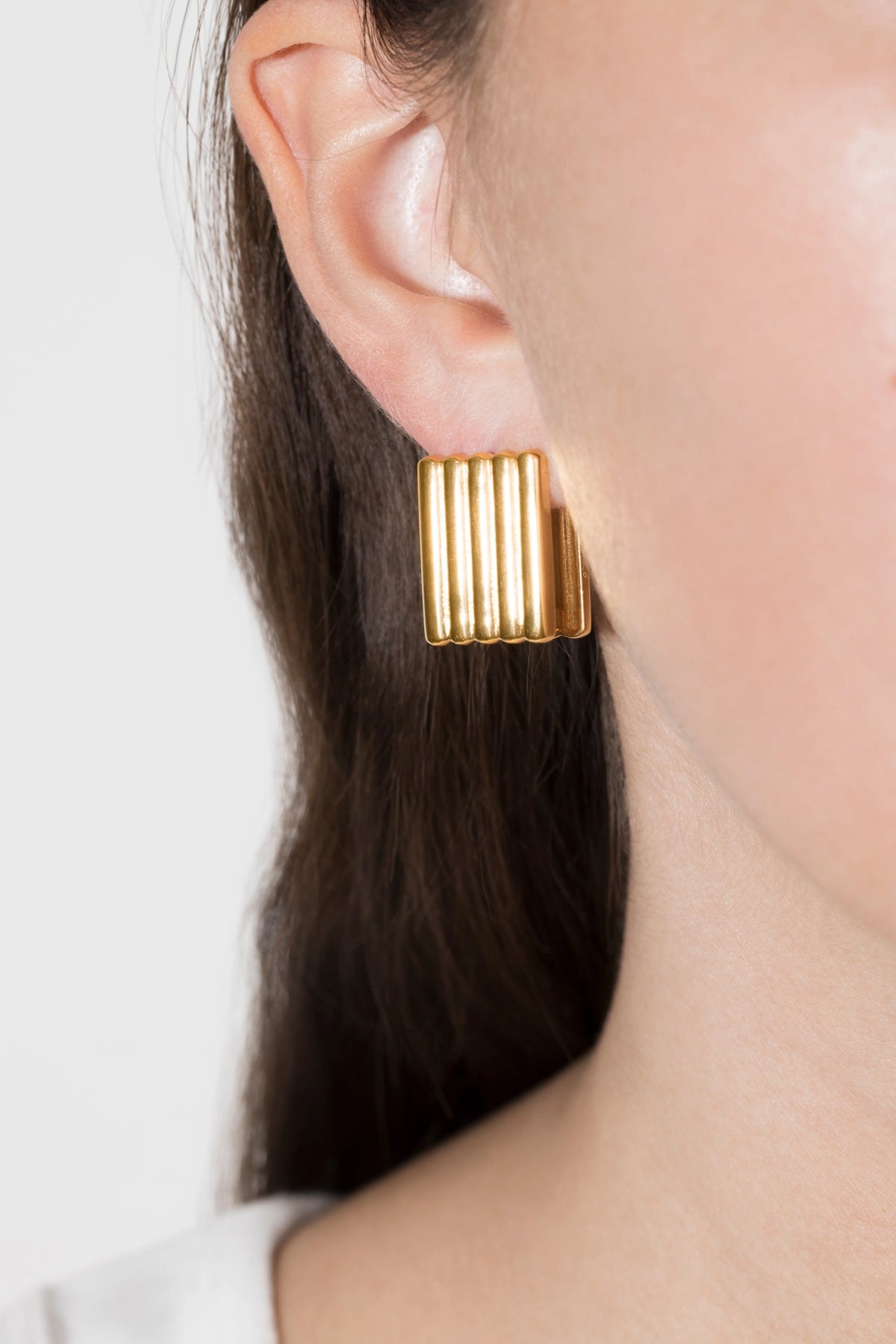 A close-up of a model's ear wearing a gold-toned, vertically ribbed rectangular stud earring against a light gray background.