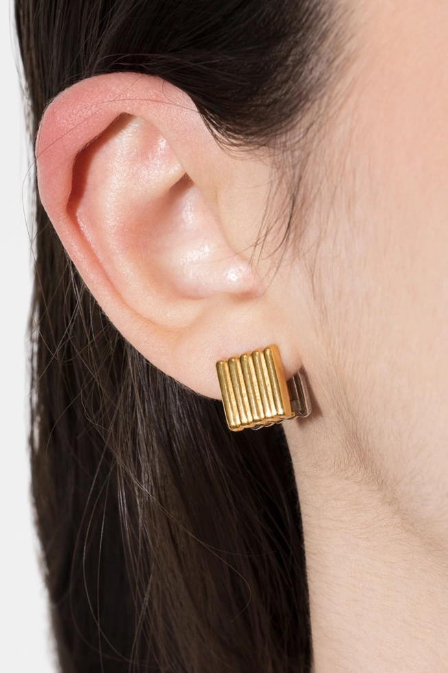 A close-up of a model's ear wearing a gold rectangular stud earring with vertical ridges, set against dark hair and a light neutral background.