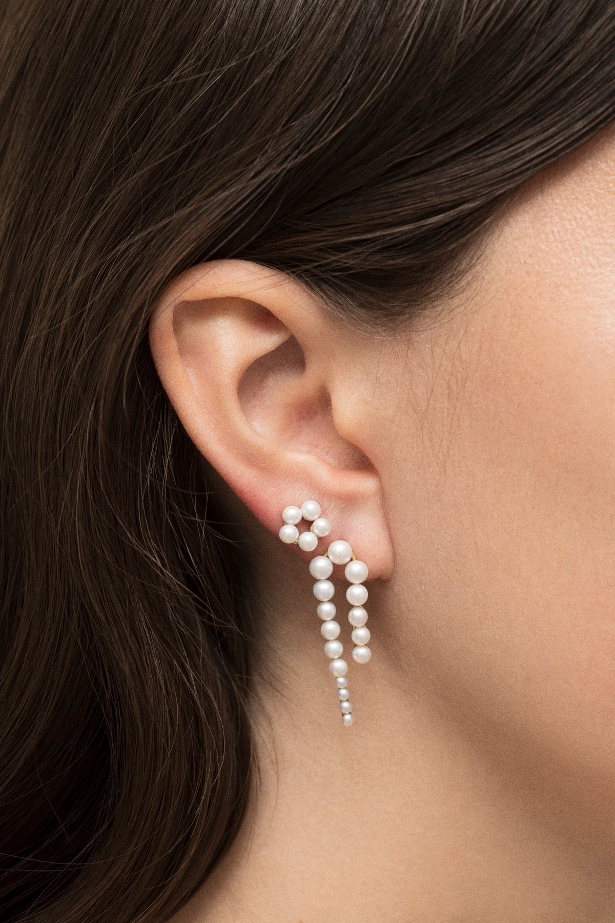 A close-up of a model's ear wearing a dangling pearl earring with multiple strands of white pearls and small clear stones.