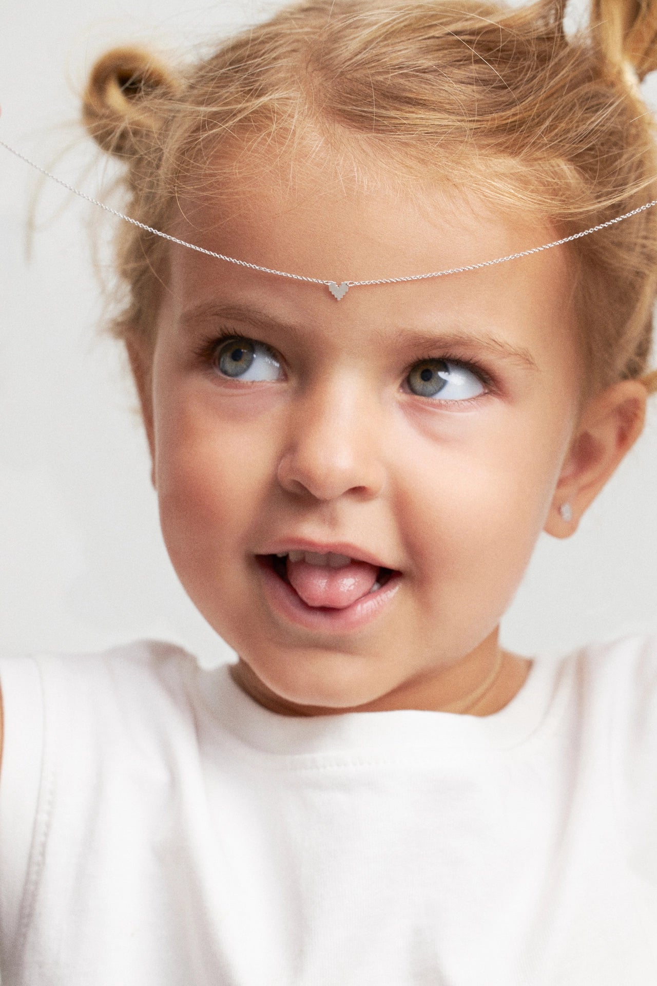 A close-up of a young child with blonde pigtails wearing a delicate silver headband with a small triangular charm, a white t-shirt, and a small stud earring, looking to the side against a light background.