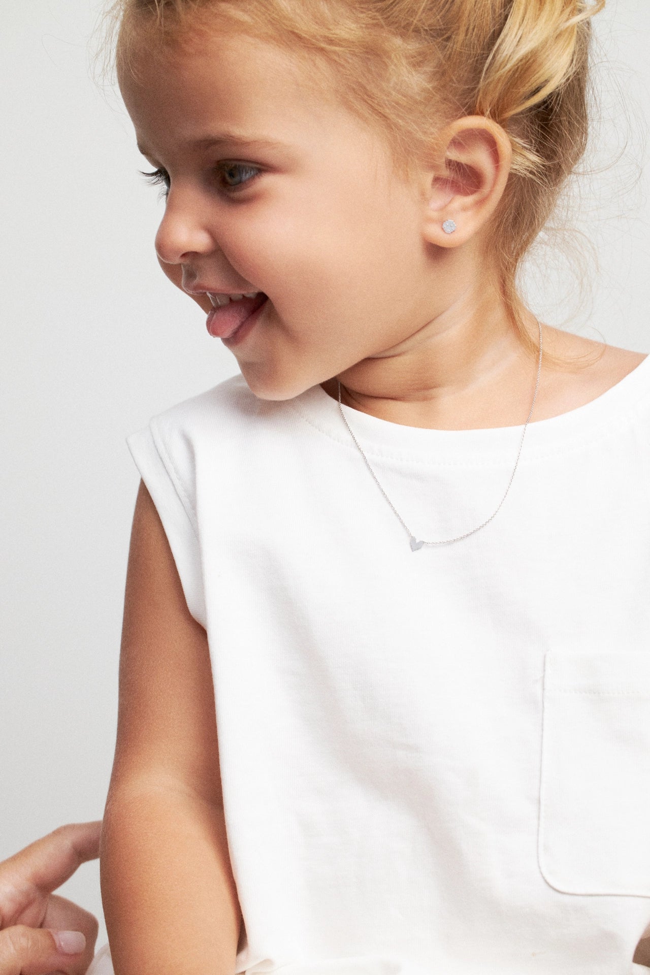 A close-up lifestyle shot of a young girl with blonde hair in a bun, wearing a white sleeveless top and a delicate silver necklace with a small heart pendant. She is looking to the side with a smile, against a clean, light gray background.