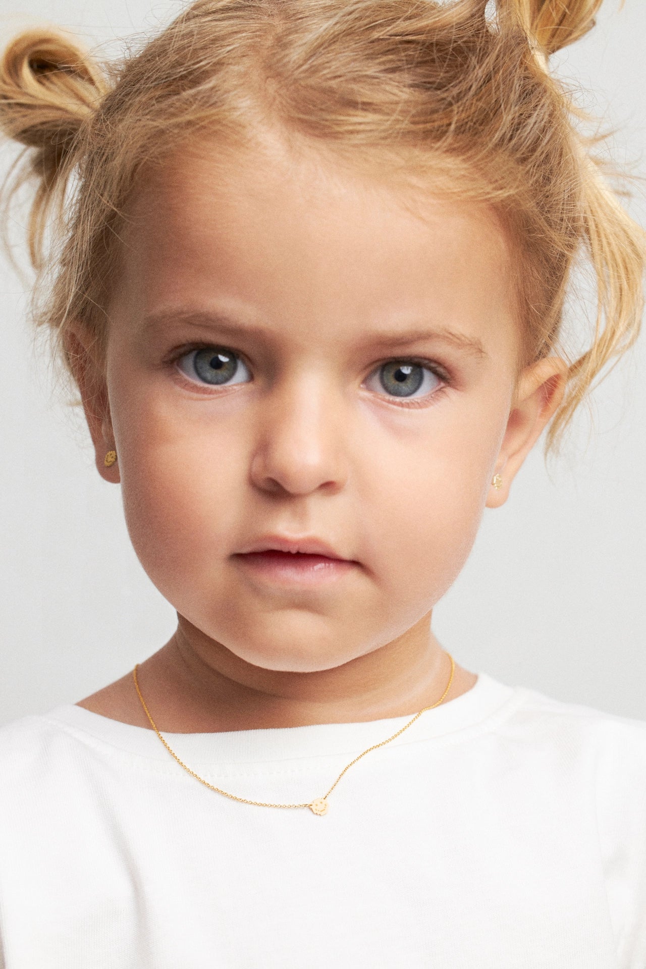 A close-up portrait of a young child with blonde pigtails wearing a delicate gold necklace and small gold earrings against a light gray background.