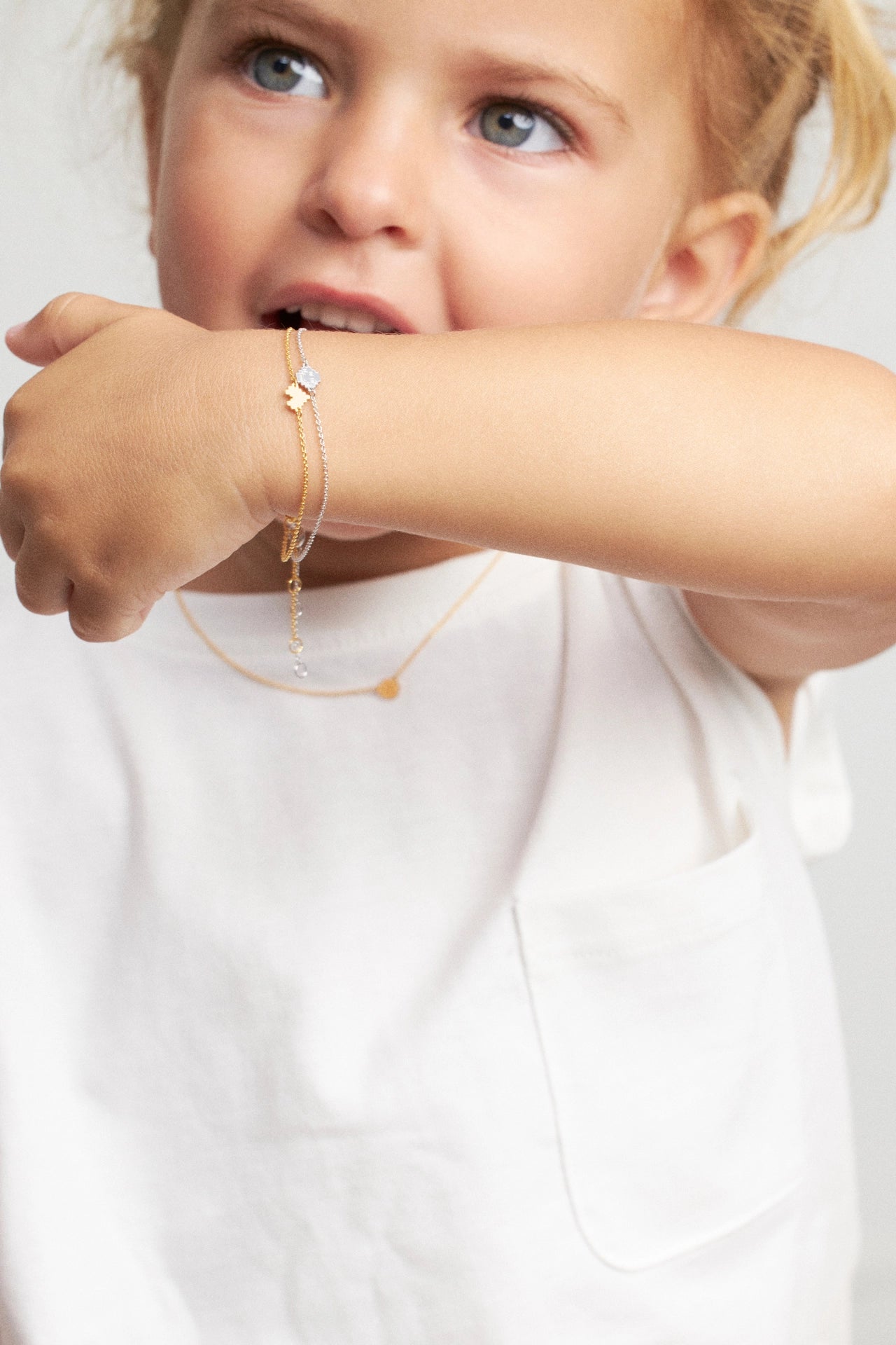 A close-up lifestyle shot of a young child wearing delicate gold jewelry, featuring a thin gold chain bracelet with a small white flower charm and a gold necklace with a tiny heart, set against a clean white background.